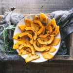 A white plate with roasted butternut squash in moon-shaped slices on a dark wood surface with gray cloth and fresh herbs.
