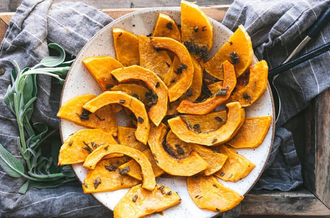 A white plate with roasted butternut squash in moon-shaped slices on a dark wood surface with gray cloth and fresh herbs.