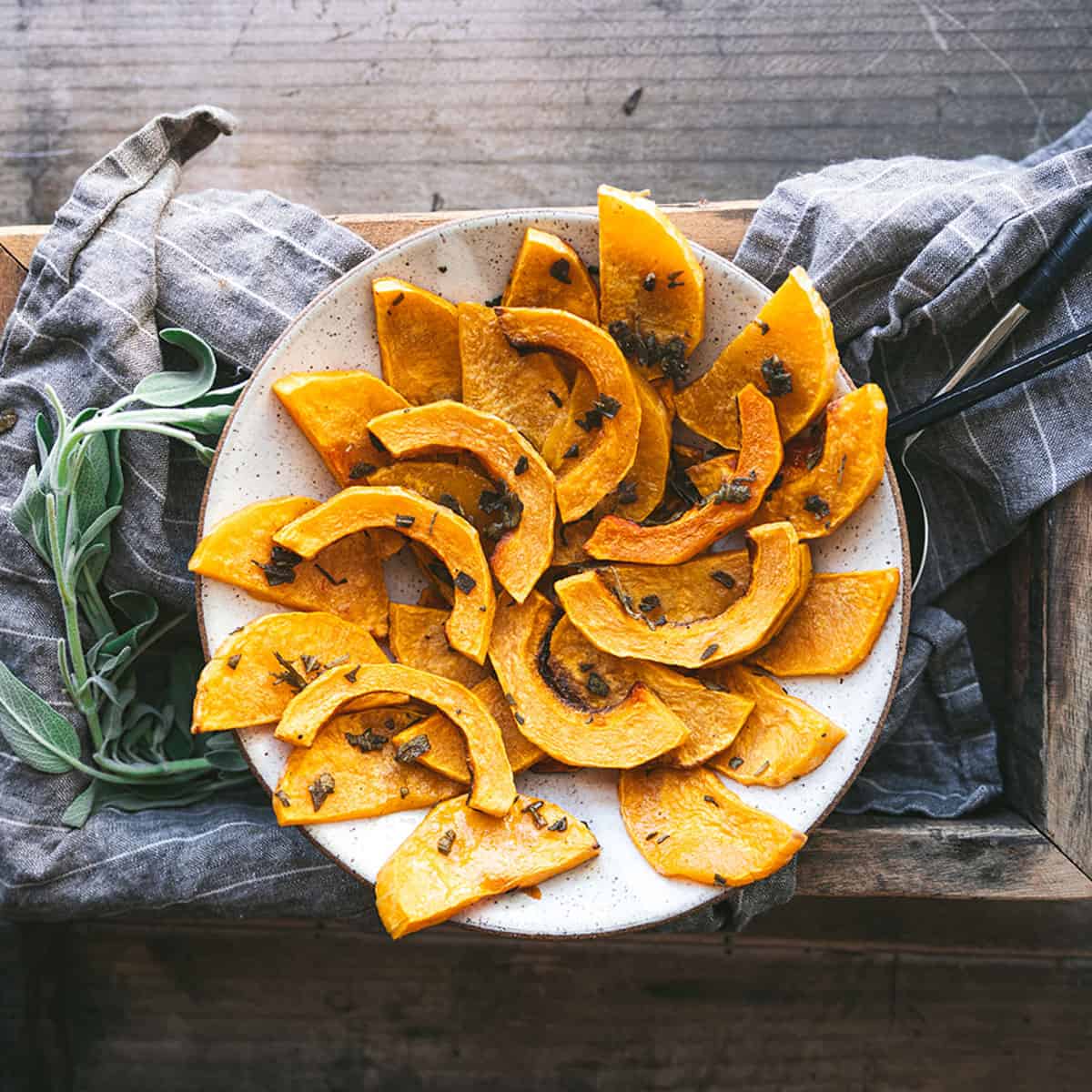 A white plate with roasted butternut squash in moon-shaped slices on a dark wood surface with gray cloth and fresh herbs. 