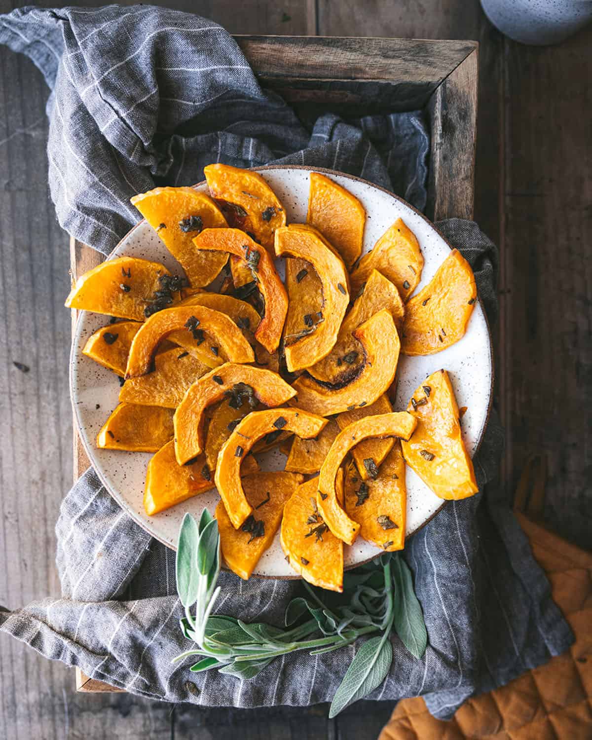Roasted butternut squash on a white plate with sage, sitting on a gray cloth on a dark wood surface. 