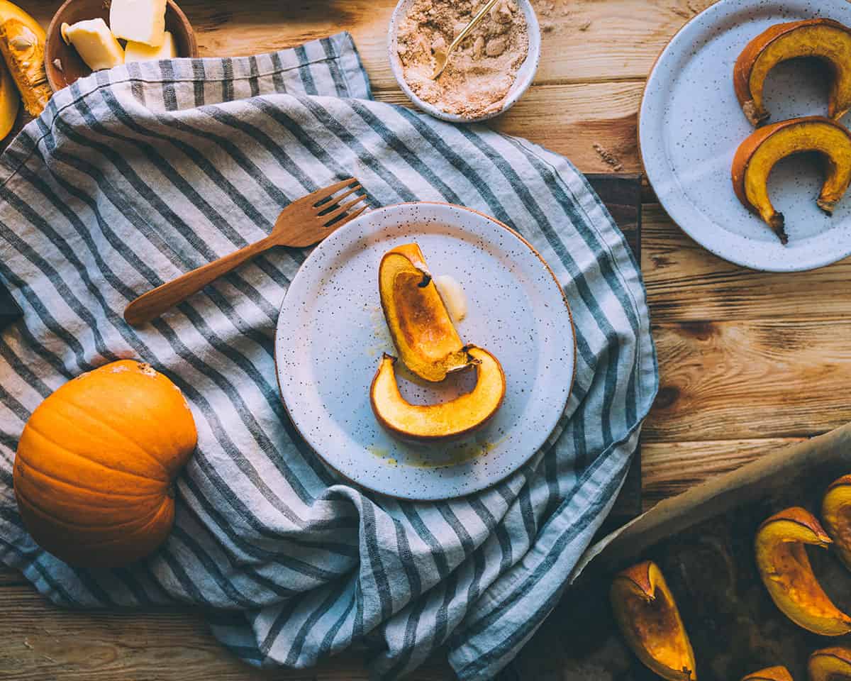 Roasted pumpkin wedges on a white plate resting on a gray and white striped towel, surrounded by other plates with pumpkin wedges, a gold fork, and ingredients for roasting pumpkins.
