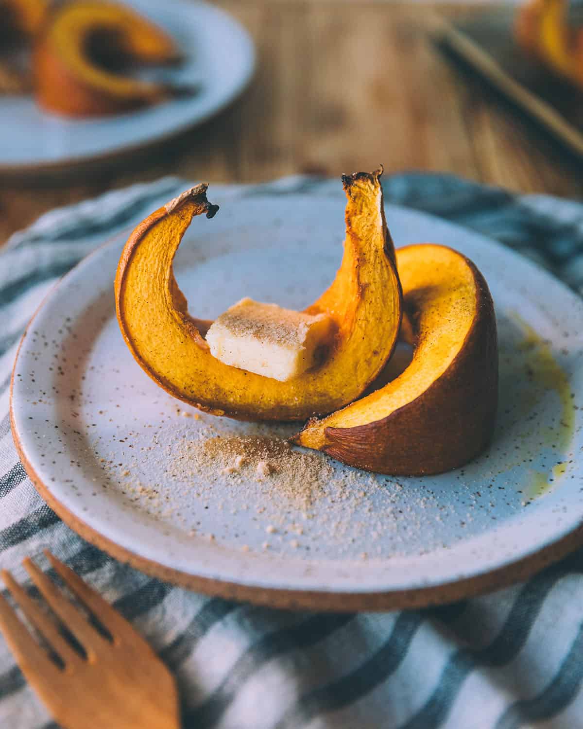 Roasted pumpkin wedges with a pat of butter on top, sitting on a white plate, with a gold fork and a grey and white towel underneath. 