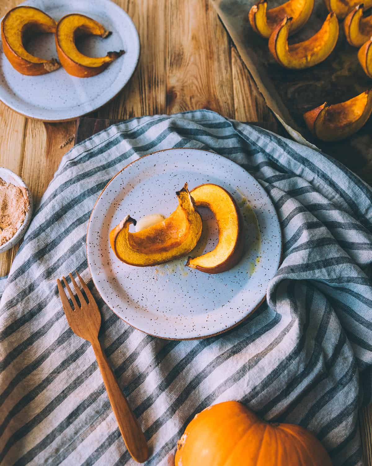 Roasted pumpkin wedges on a white plate resting on a gray and white striped towel, surrounded by other plates with pumpkin wedges, a gold fork, and ingredients.