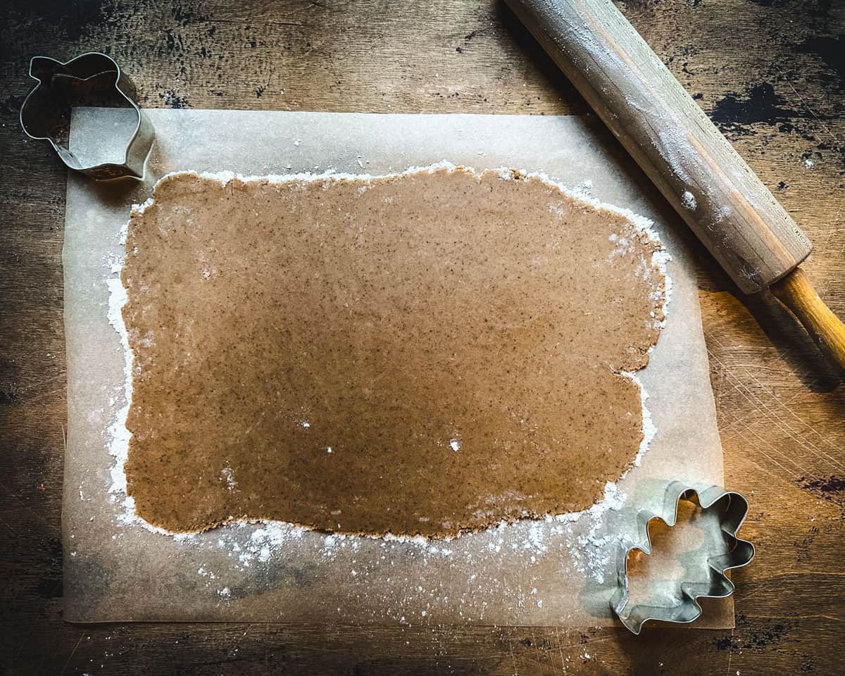 Acorn flour dough rolled out on parchment paper sitting on a wood surface, surrounded by a wood rolling pin and cookie cutters. 