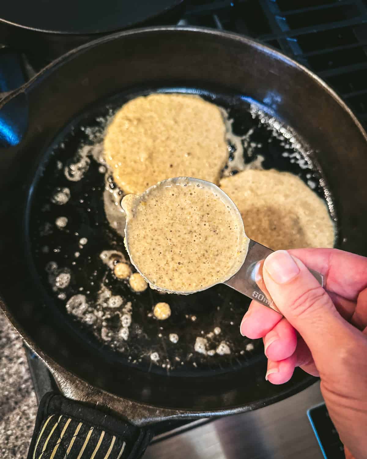 A measuring cup pouring acorn pancake batter into a cast iron skillet. 