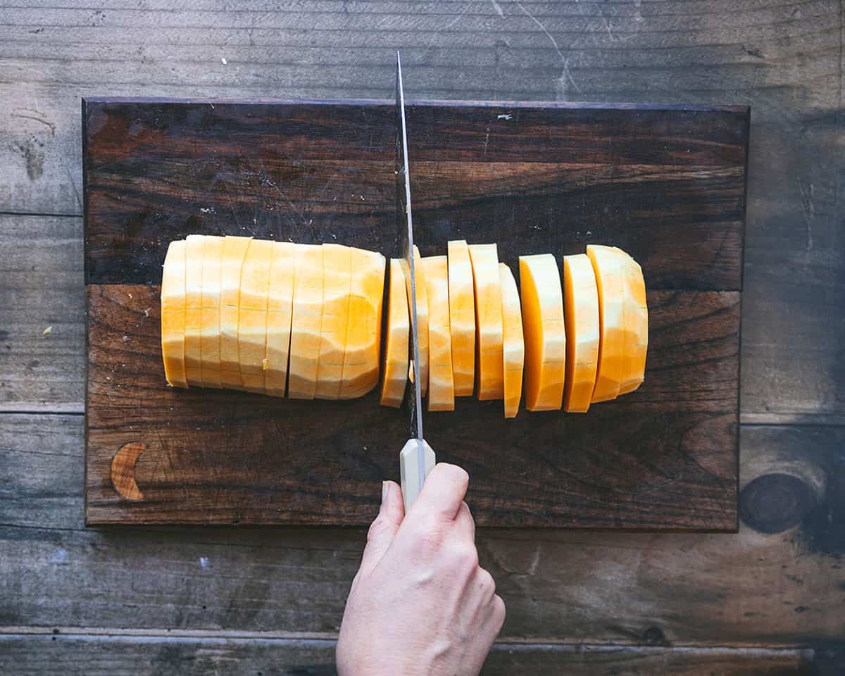 Butternut squash being sliced with flat side down on a dark wood cutting board. 