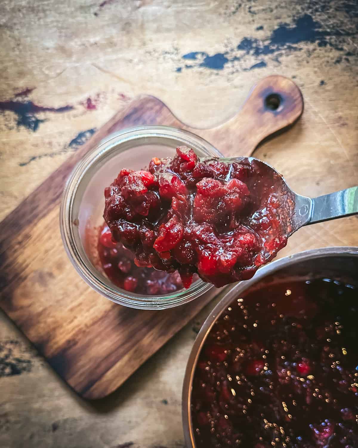 Cooked cranberries being spooned into a pickling jar, sitting on a wood cutting board. 