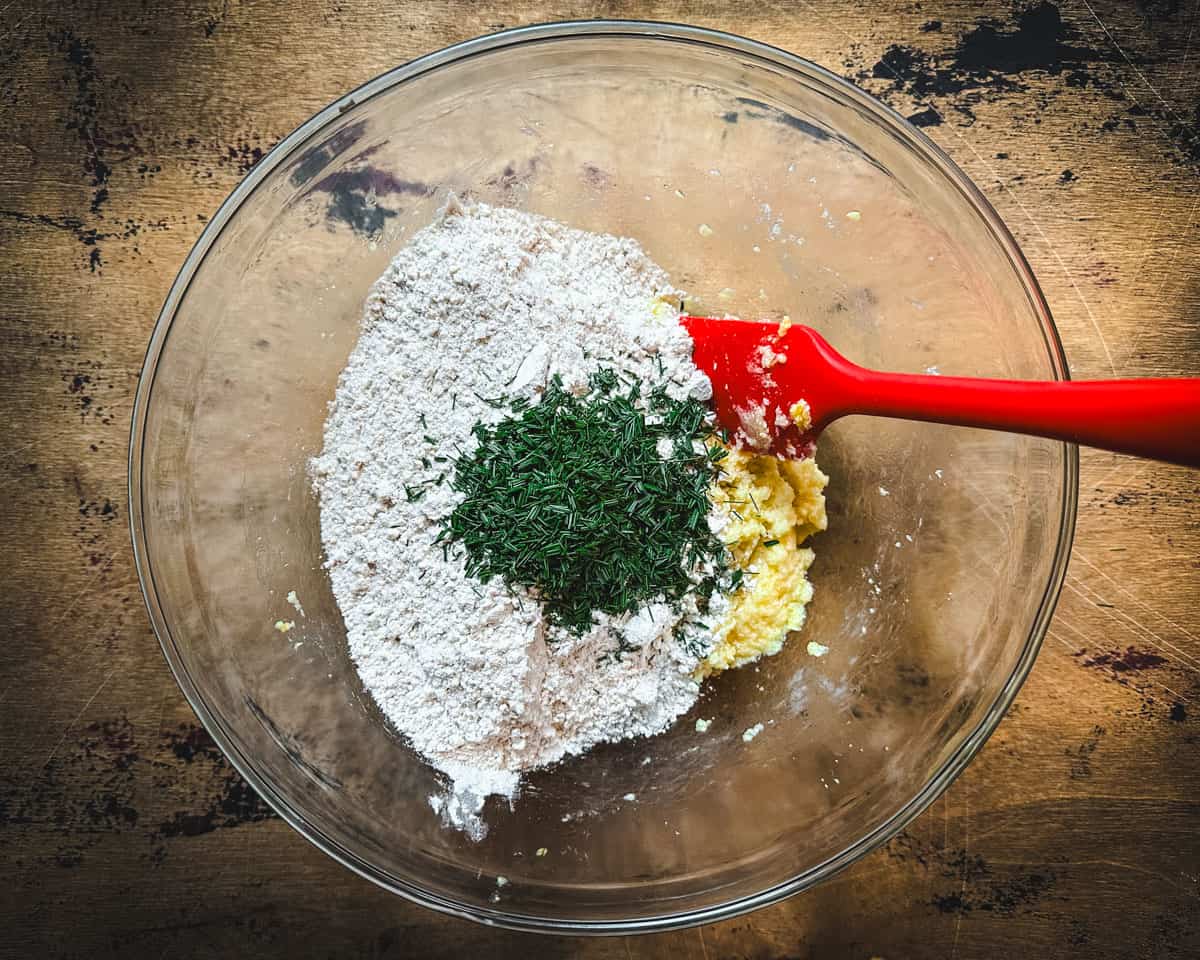 Dry ingredients added to the creamed butter with pine needles on top in a clear bowl on a wood surface. 