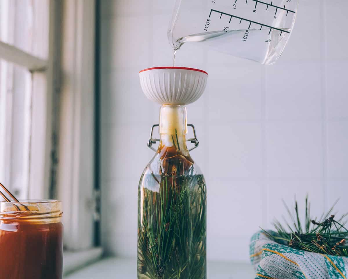 Water pouring from a glass measuring cup into a bottle with pine needles and honey, with a funnel on top. Surrounded by a jar of honey, and a bowl of pine needles.