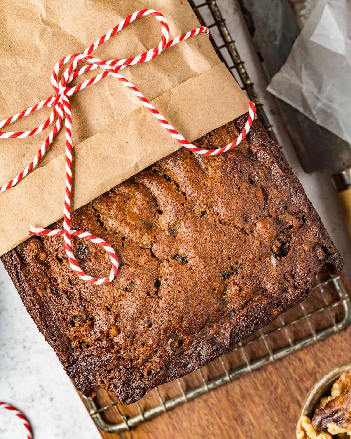 Fruit cake on a cooling rack, half covered with brown parchment paper and tied with red and white twine, sitting on a wood surface. 