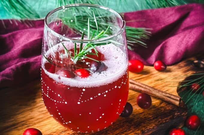 A wine glass with white constellations on it filled halfway with cranberry champagne cocktail and garnished with cranberries and rosemary. On a wood surface, surrounded by fresh cranberries, green and red cloths, and pine.