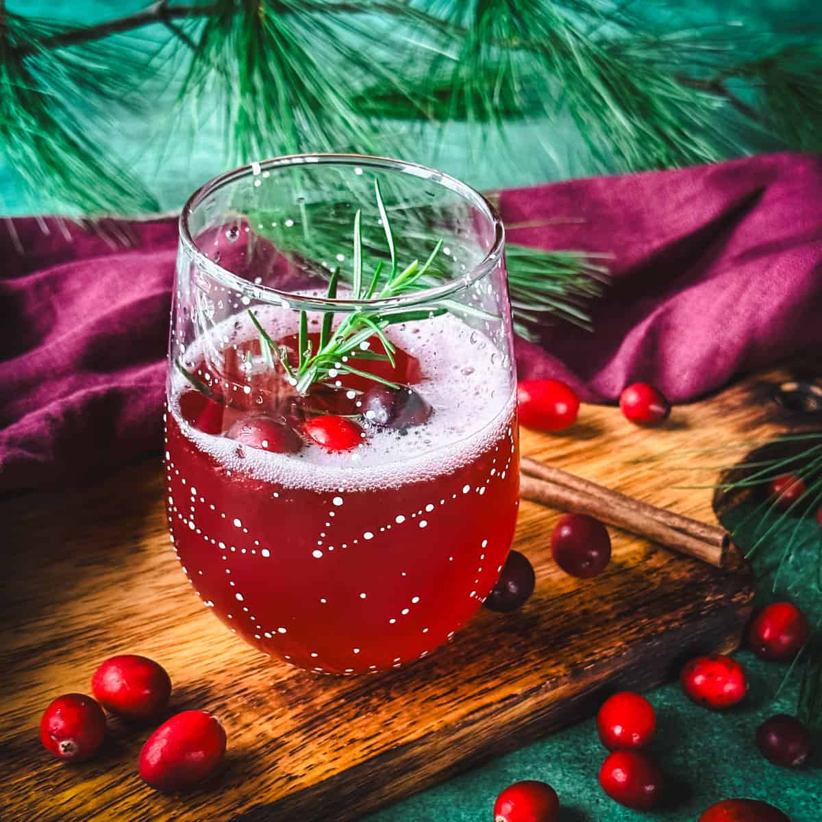 A wine glass with white constellations on it filled halfway with cranberry champagne cocktail and garnished with cranberries and rosemary. On a wood surface, surrounded by fresh cranberries, green and red cloths, and pine. 