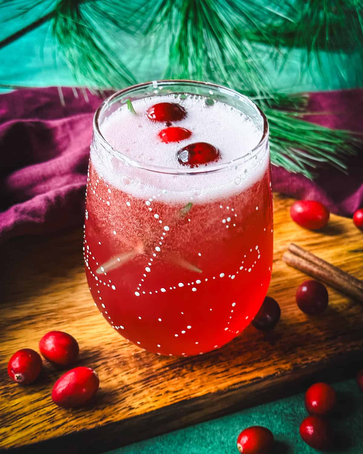 A wine glass with white constellations on it filled with cranberry champagne cocktail and garnished with cranberries and rosemary. On a wood surface, surrounded by fresh cranberries, green and red cloths, and pine.