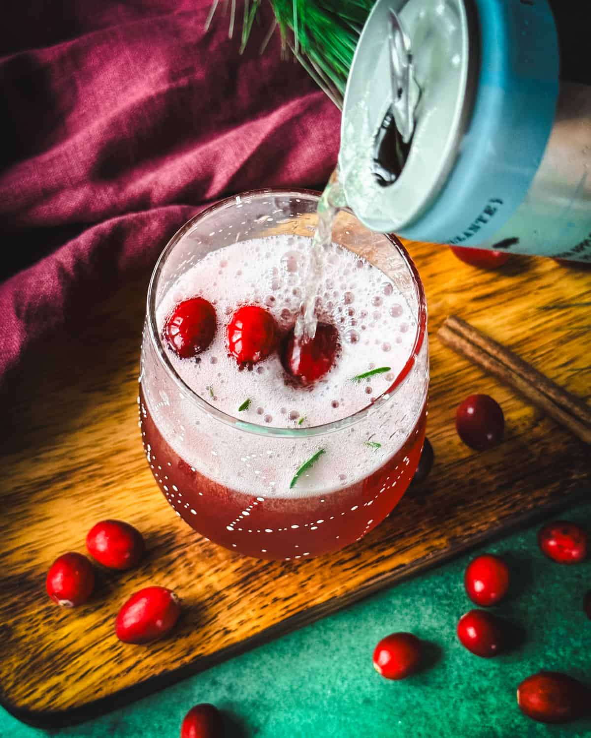 A stemless wine glass with cranberry champagne cocktail being topped off with more sparkling wine from a can. On a wood surface with a red cloth and fresh cranberries surrounding. 