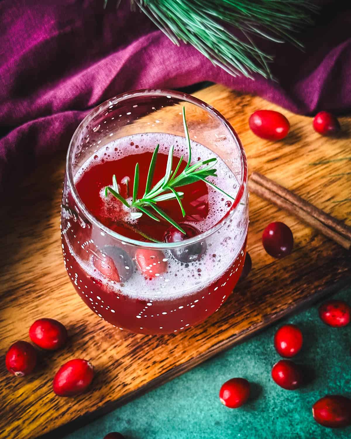 A cranberry champagne cocktail garnished with whole cranberries and rosemary, top view. On a wood and green surface, surrounded by fresh cranberries and a red cloth. 