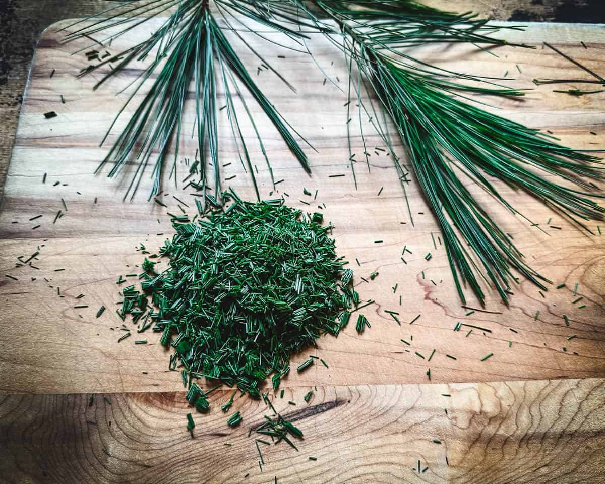 Finely chopped pine needles on a wood cutting board, surrounded by pine needles. 