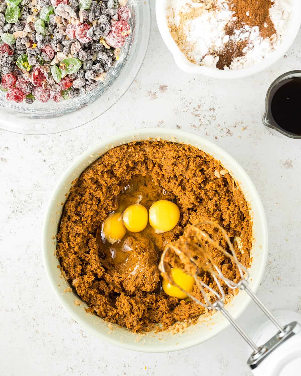 A white mixing bowl with cake batter that is brown from molasses, and raw eggs with a whisk attachment to beat it in. 