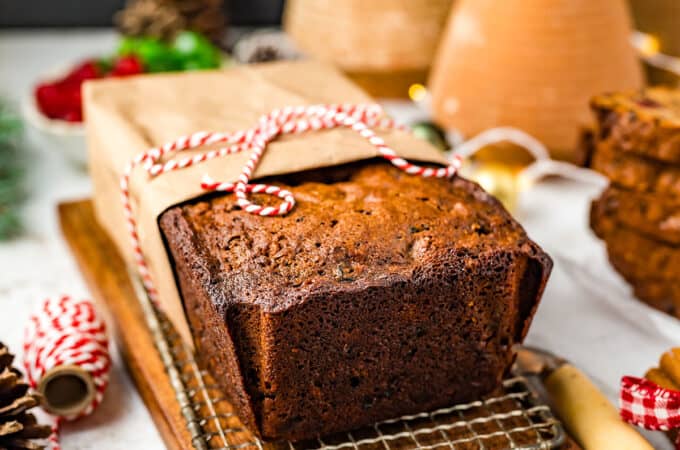 A fruit cake half-wrapped in brown parchment and tied with red and white string, on a cooling rack resting on a cutting board. Surrounded by string and other holiday decorations.