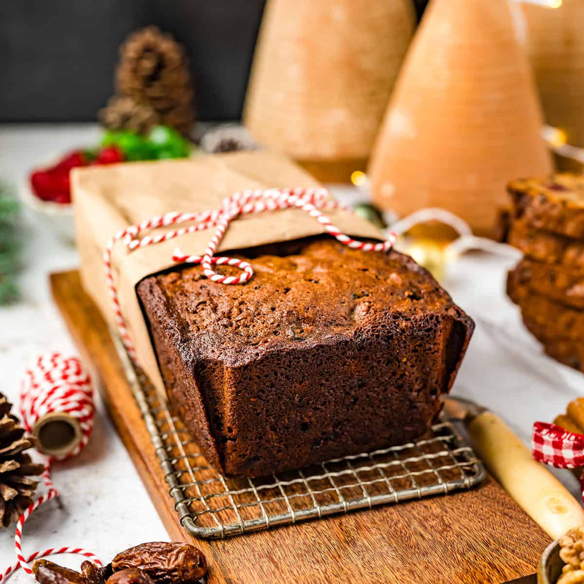 A fruit cake half-wrapped in brown parchment and tied with red and white string, on a cooling rack resting on a cutting board. Surrounded by string and other holiday decorations. 