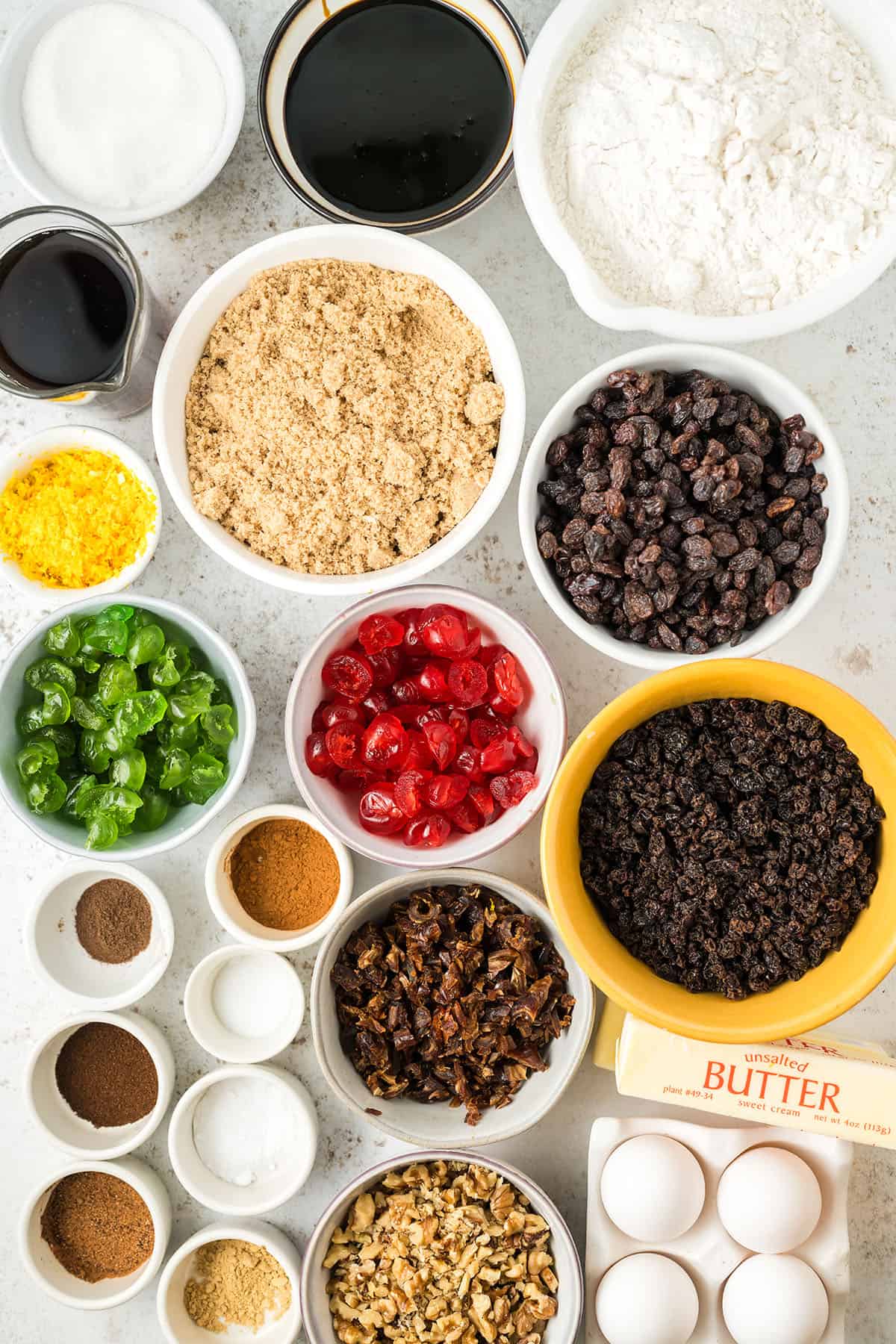 Many bowls of ingredients for fruit cake, including dried and candied fruit, flour, sugar, and molasses. 