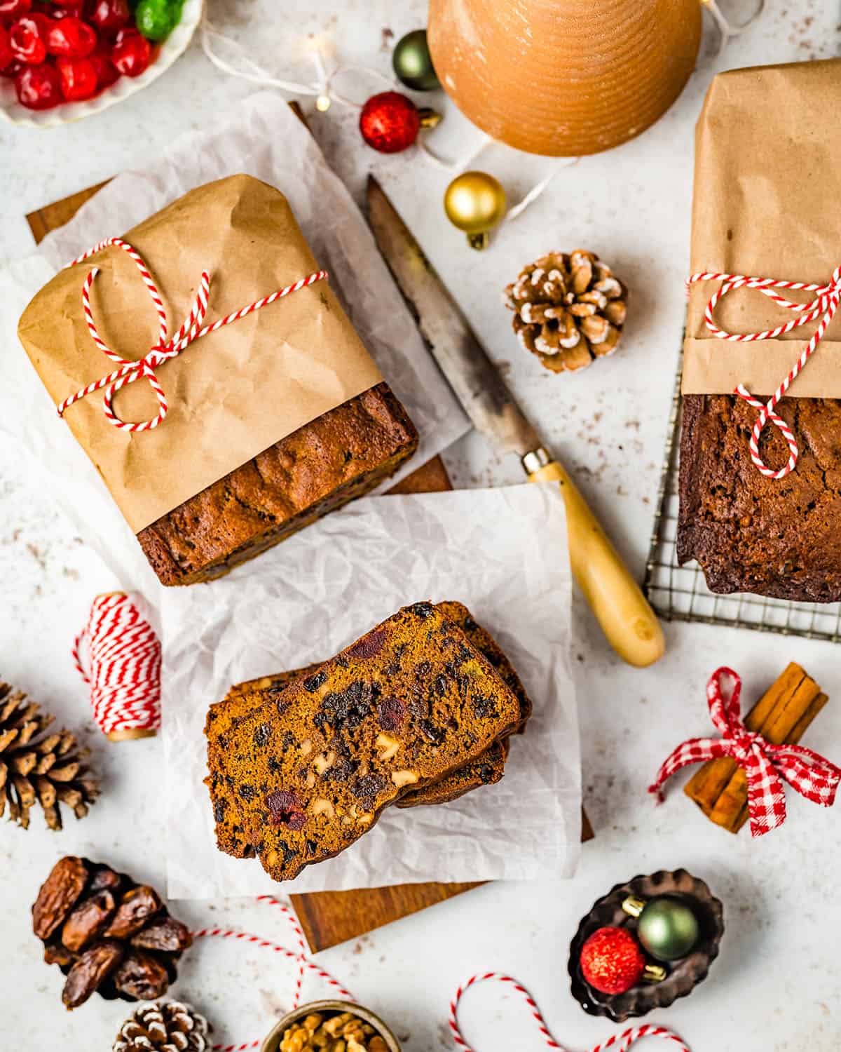 A loaf of fruit cake with half in slices, while the other half is wrapped in parchment paper tied with red and white twine. Surrounded by a loaf of fruit cake still in the pan, pine cones, and small ornaments. 