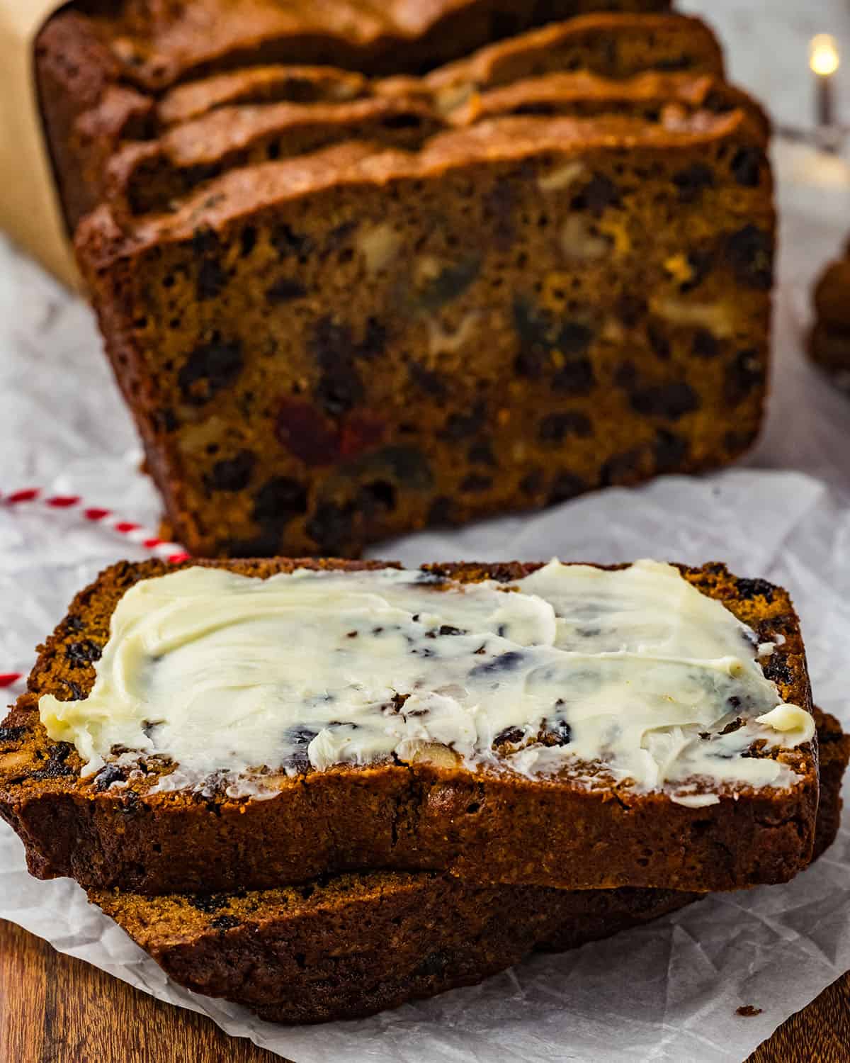 A fruit cake cut into slices, with two laying face up on a white parchment with a thick layer of butter on the top slice. 