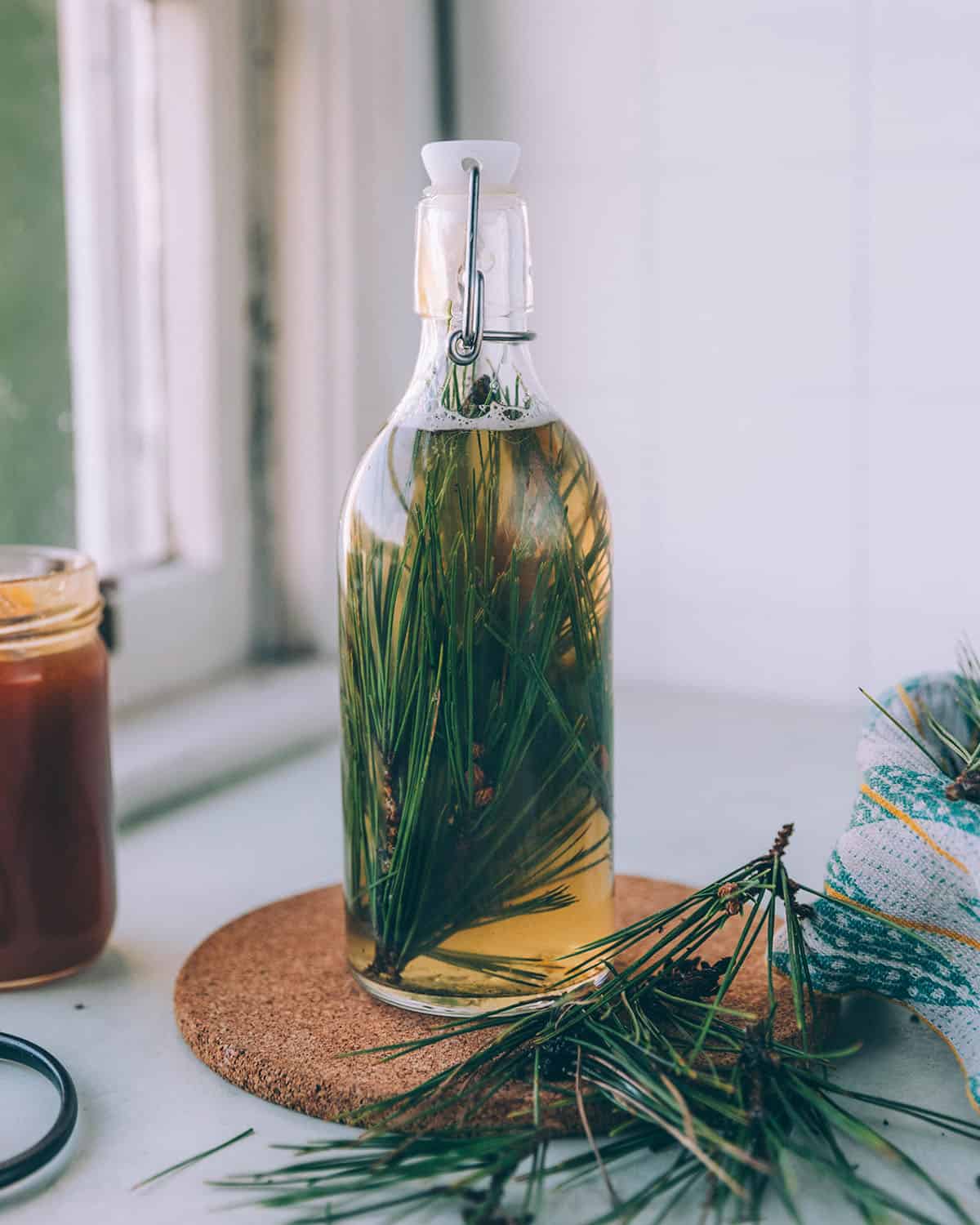 A bottle of pine needle soda ready to ferment, on a circular trivet, sitting on a white counter surrounded by a jar of honey, and pine needles. 
