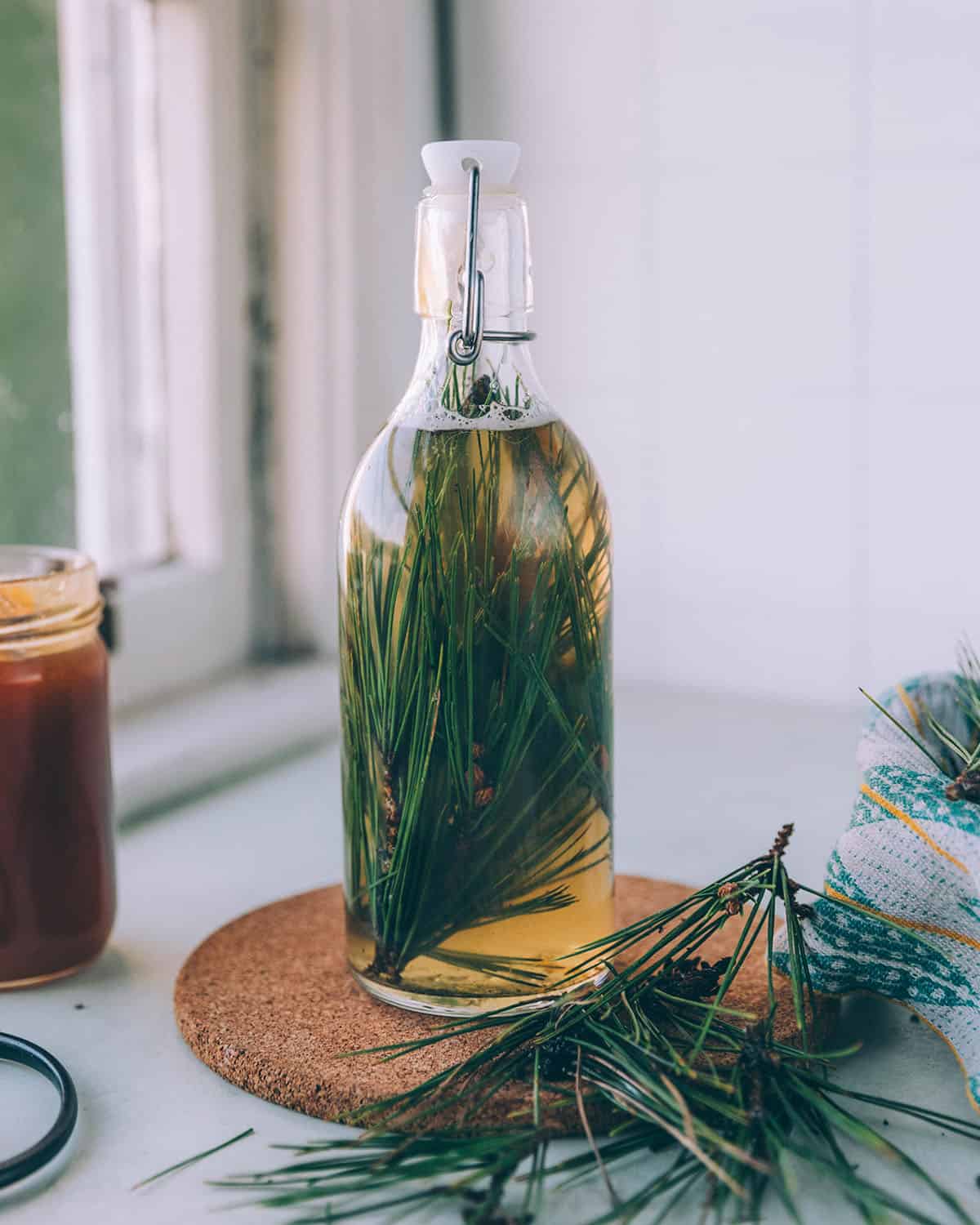 A bottle of pine needle soda ready to ferment, on a circular trivet, sitting on a white counter surrounded by a jar of honey, and pine needles.