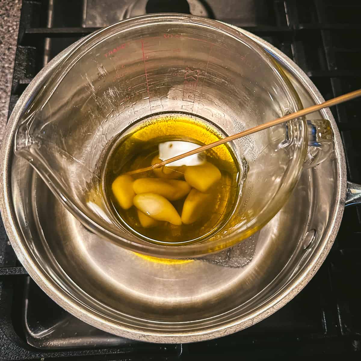 A makeshift double boiler with melted oils and melting beeswax being stirred with a bamboo skewer, top view. 