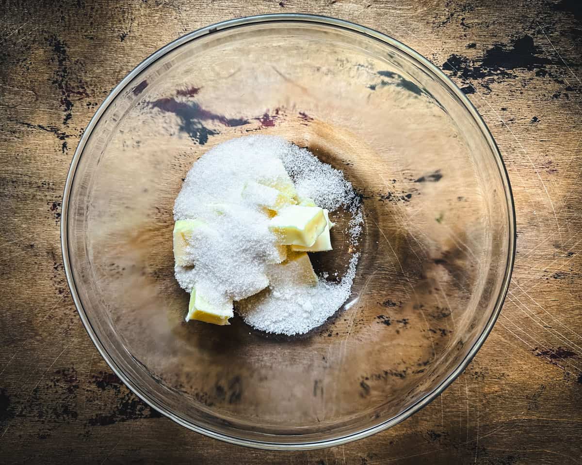 A clear bowl with butter and sugar, on a dark wood surface, top view. 