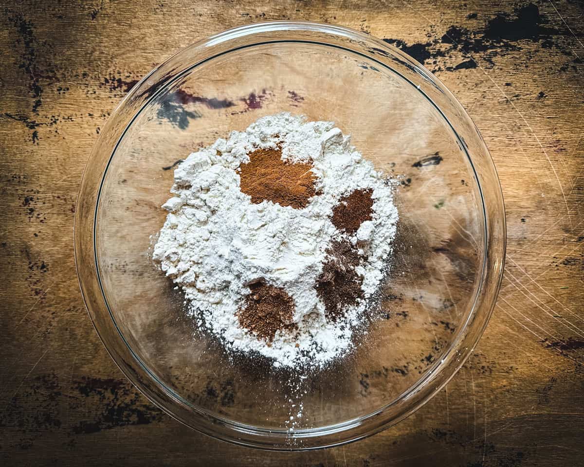 A clear bowl with dry cookie ingredients, on a dark wood surface. 