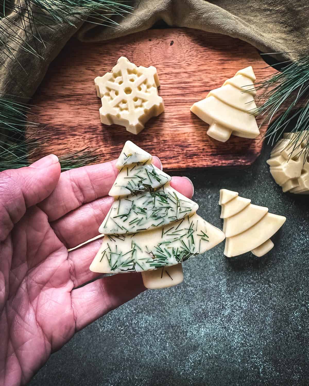 Cream colored pine lotion bars on a wood board and a green counter top, with a hand holding one up that is tree shaped with pine needles decorating it. 