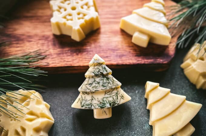 Christmas tree and snowflake shaped lotion bars, cream colored. One tree has pine needles sprinkled on it, on a wood cutting board and a dark green surface, with pine needles surrounding.