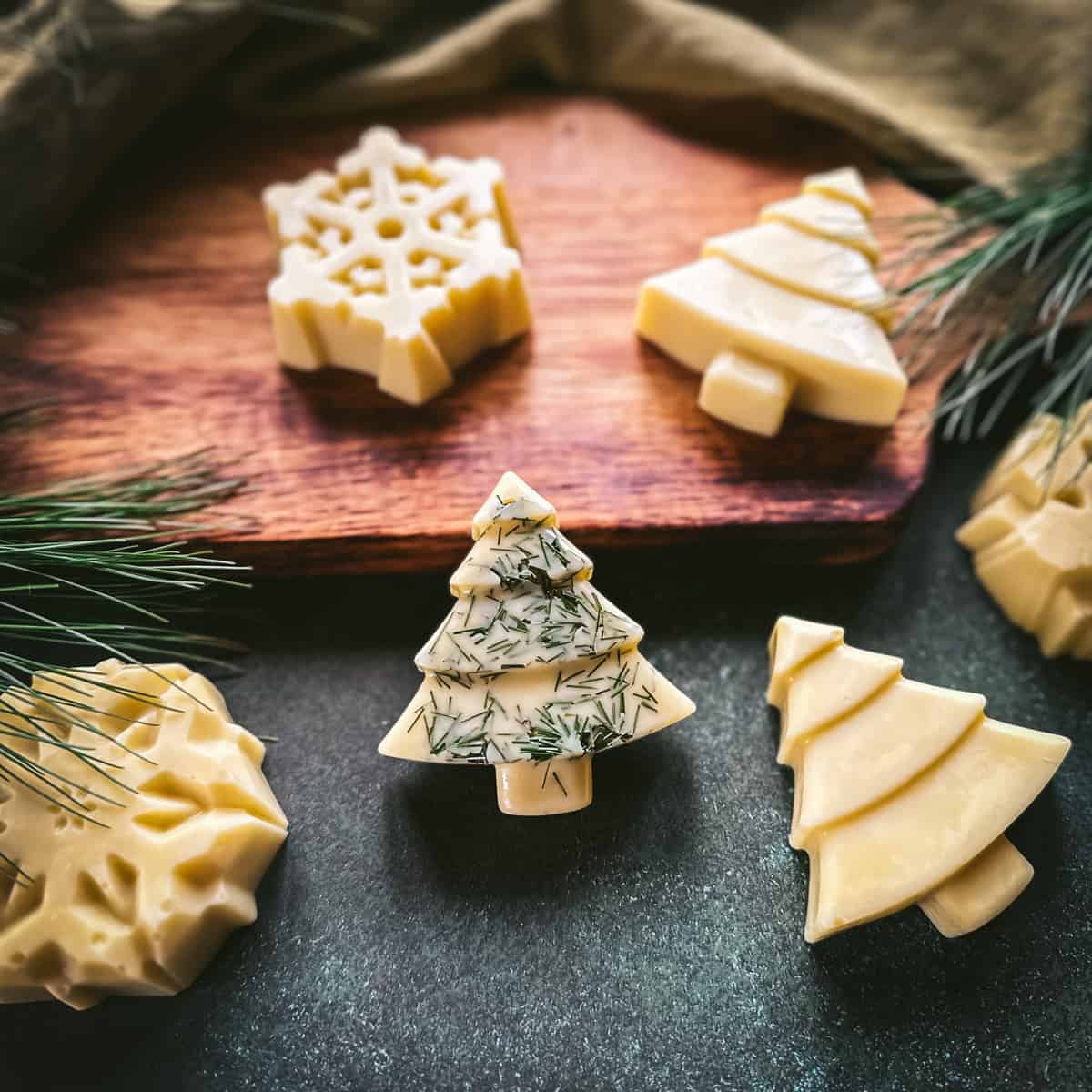 Christmas tree and snowflake shaped lotion bars, cream colored. One tree has pine needles sprinkled on it, on a wood cutting board and a dark green surface, with pine needles surrounding.