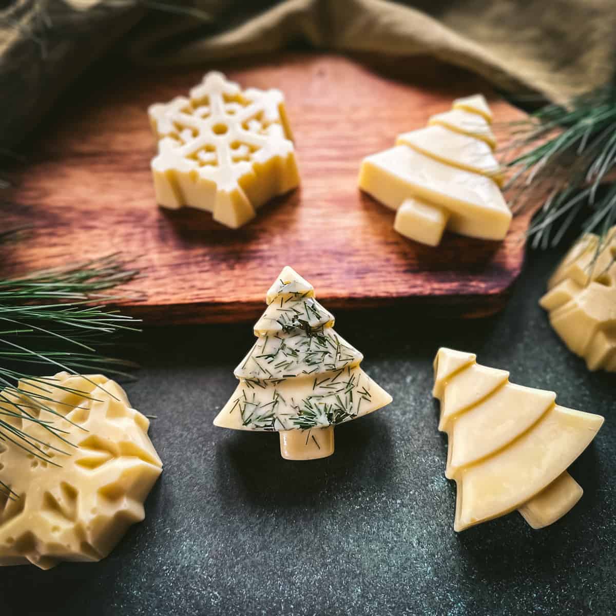 Christmas tree and snowflake shaped lotion bars, cream colored. One tree has pine needles sprinkled on it, on a wood cutting board and a dark green surface, with pine needles surrounding. 