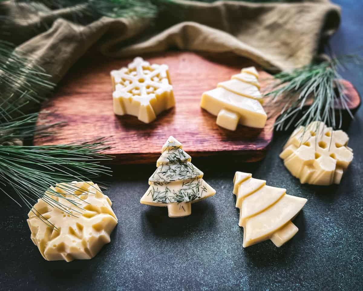 Christmas tree and snowflake shaped lotion bars, cream colored. One tree has pine needles sprinkled on it, on a wood cutting board and a dark green surface, with pine needles surrounding.