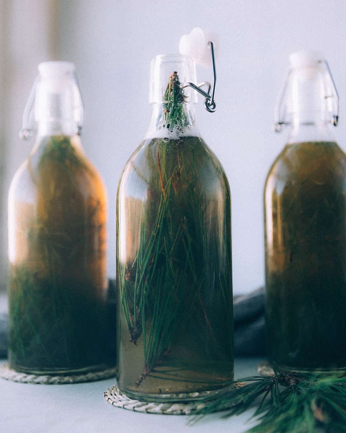 3 bottles of fermented pine needle soda with the needles still in them, on a white surface surrounded by pine needles.