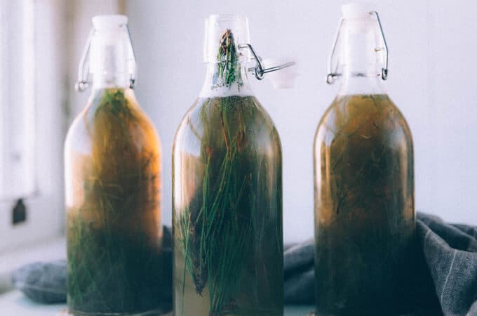 3 bottles of fermented pine needle soda with the needles still in them, on a white surface surrounded by pine needles.