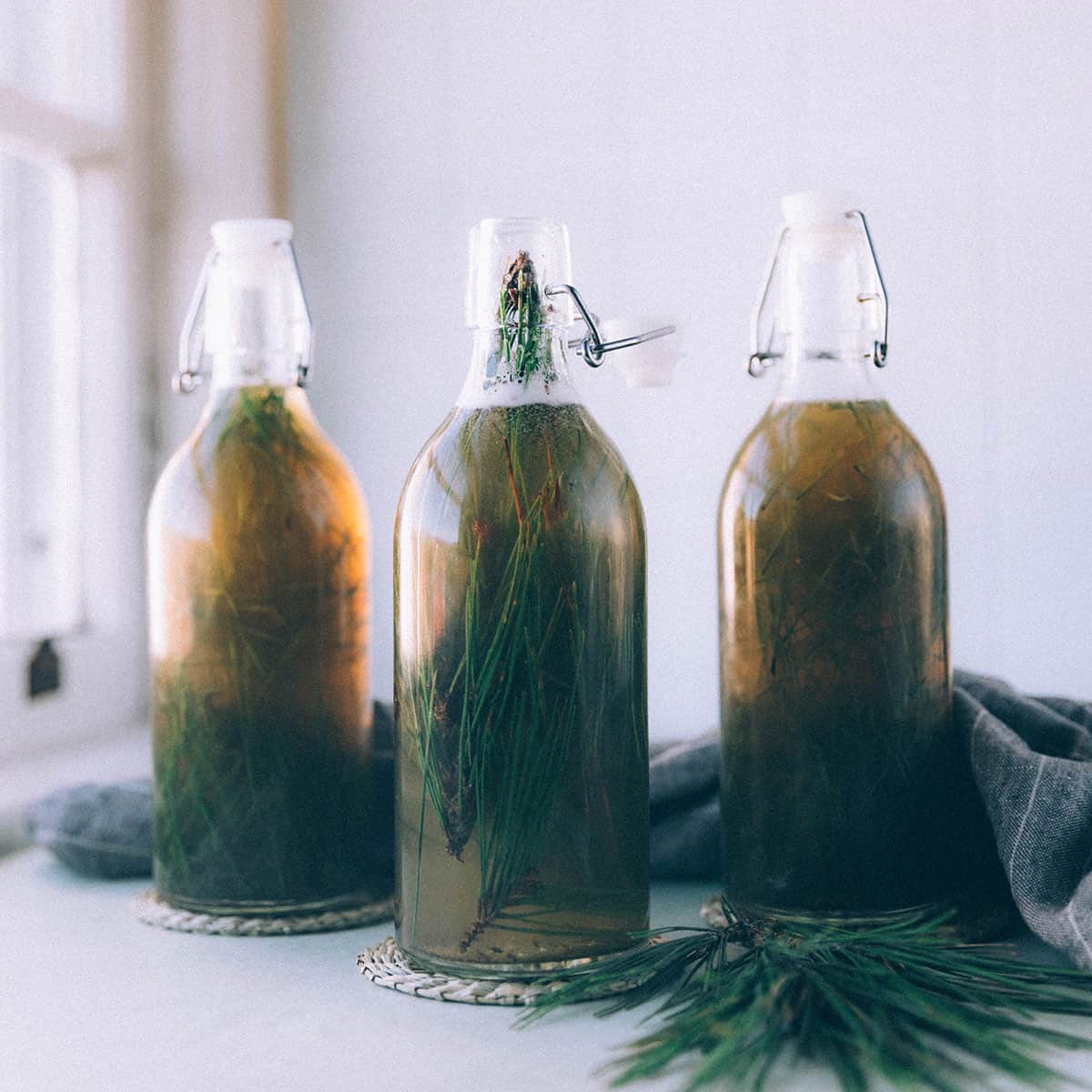 3 bottles of fermented pine needle soda with the needles still in them, on a white surface surrounded by pine needles.