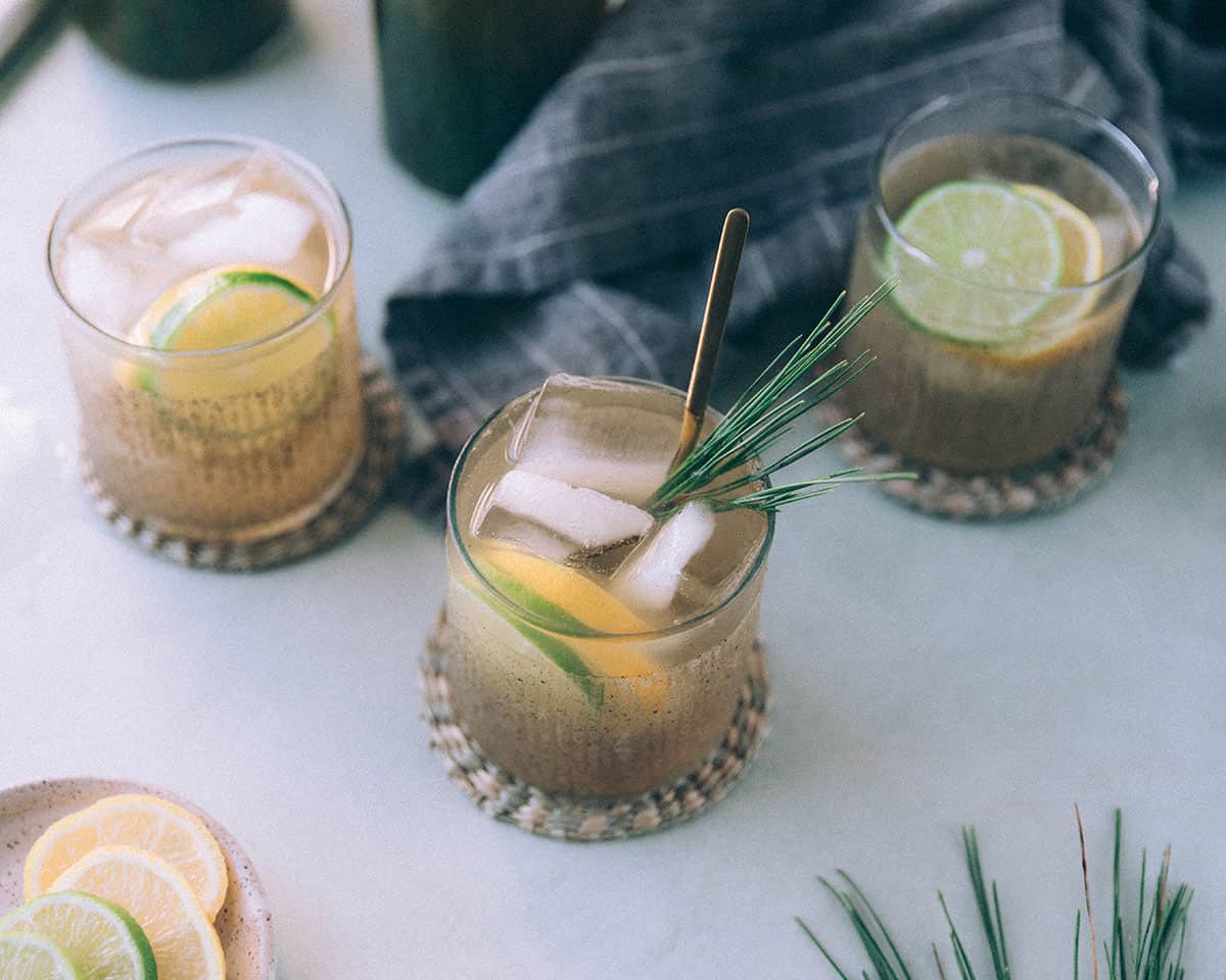 3 collins glasses with pine needle soda and garnished with lemon and lime slices, and fresh pine needles, surrounded by a gray towel and fresh pine needles.