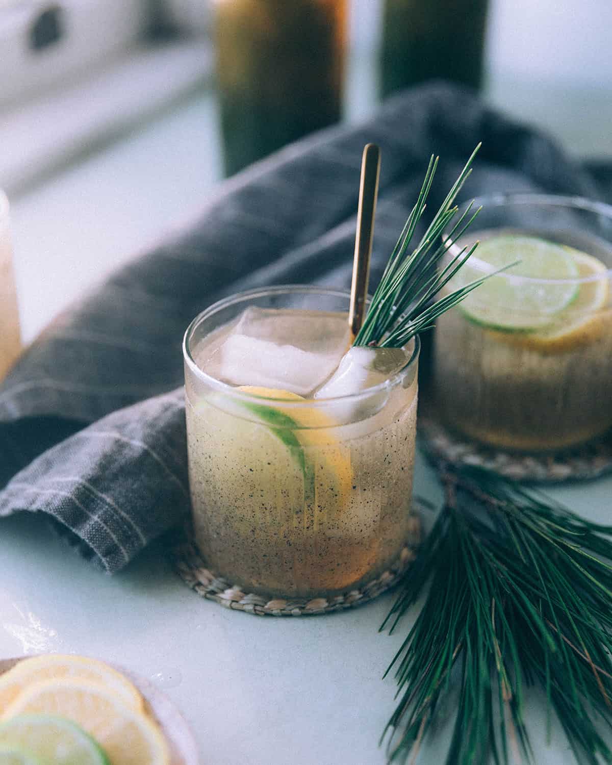 A glass of pine needle soda on ice garnished with lemon and lime slices, fresh pine needles, and a gray towel.