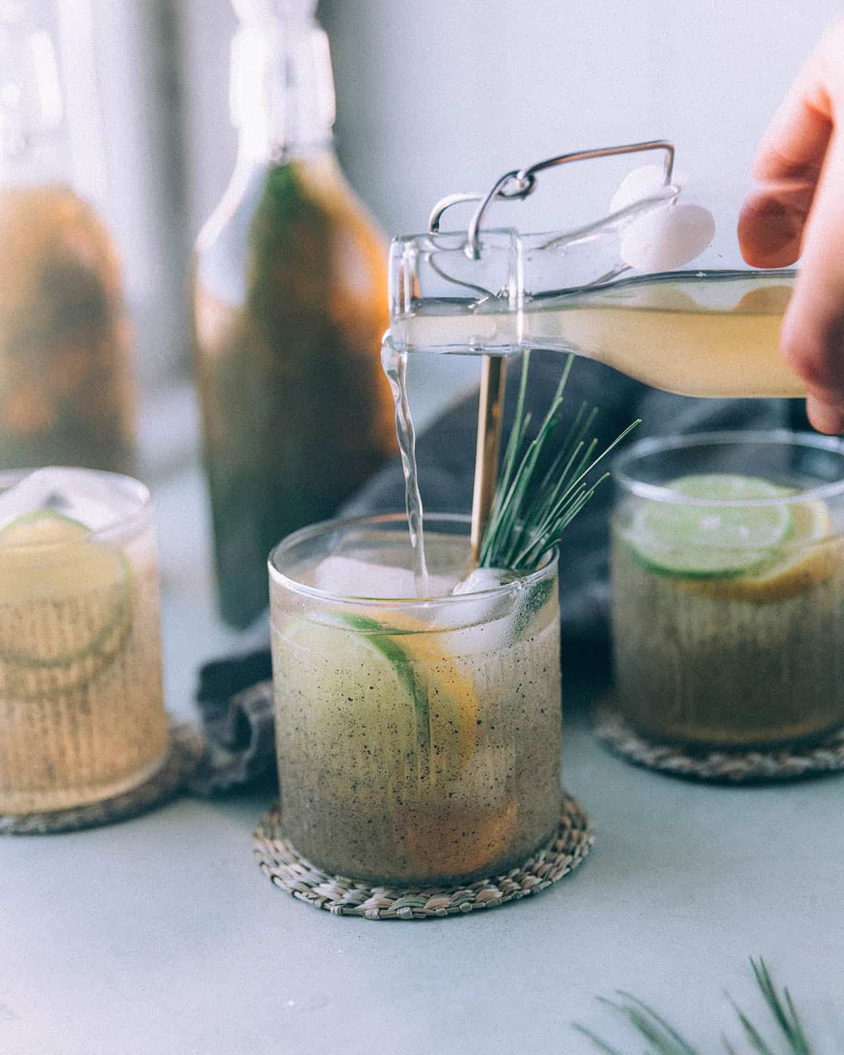 Pine needle soda pouring from a bottle into a collins glass over ice and garnished with lemon and lime slices. 