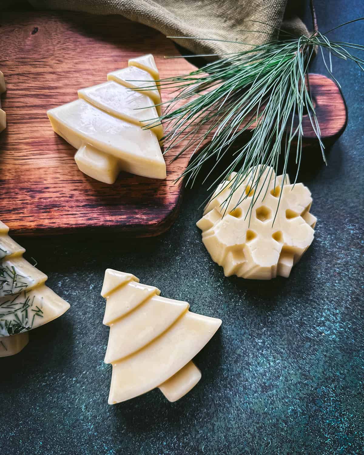 Christmas tree and snowflake shaped lotion bars, cream colored. One tree has pine needles sprinkled on it, on a wood cutting board and a dark green surface, with pine needles surrounding.