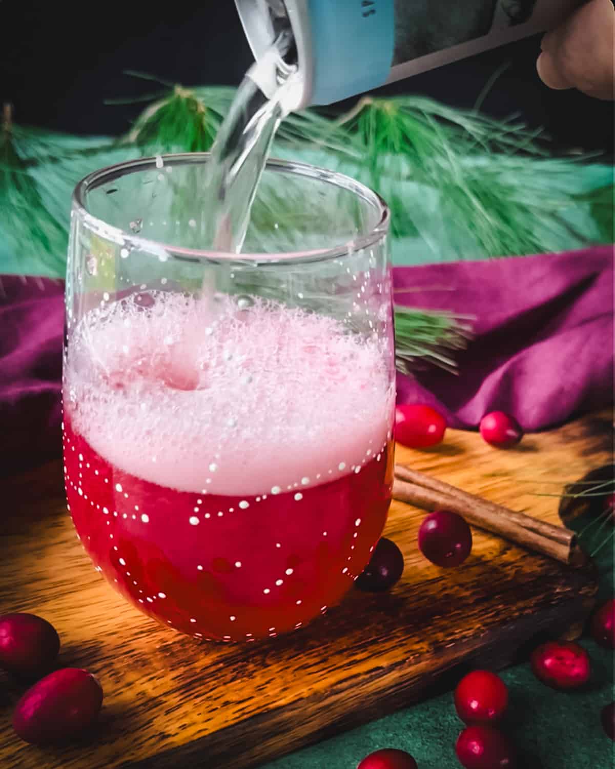 A can of champagne pouring into the wine glass over the cranberry syrup, on a wood surface surrounded by red and green cloths and fresh whole cranberries. 
