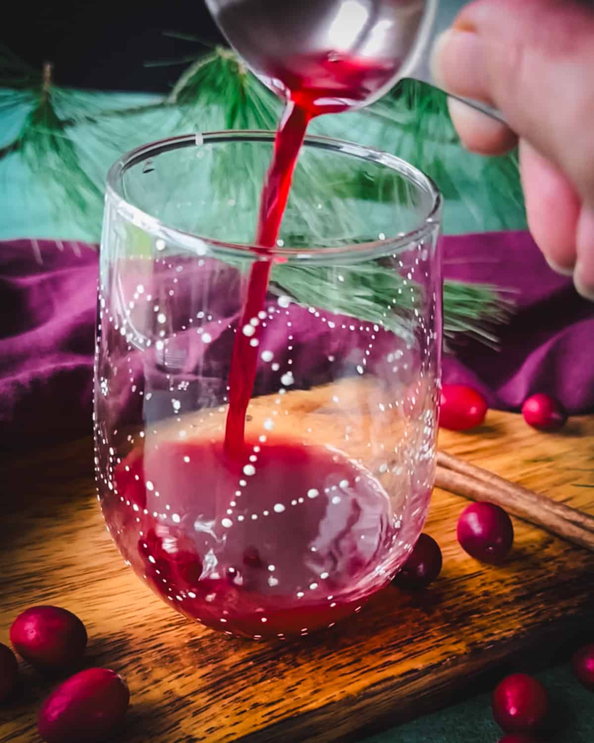 Finished spiced cranberry syrup pouring into a stemless wine glass with constellations on it, on a wood surface surrounded by red and green cloths and fresh whole cranberries. 