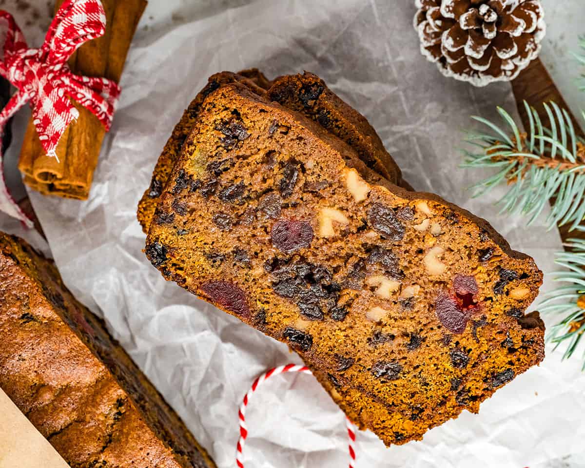 Slices of fruit cake face up on white parchment paper surrounded by red and white twine, evergreen sprigs, pinecones, and cinnamon sticks. 