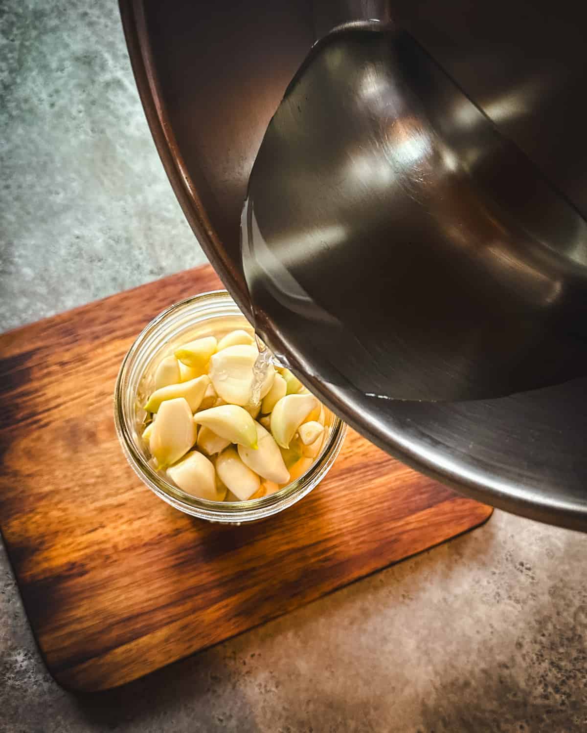 Brine pouring from a pot into a jar with garlic cloves in it on a wood cutting board. 