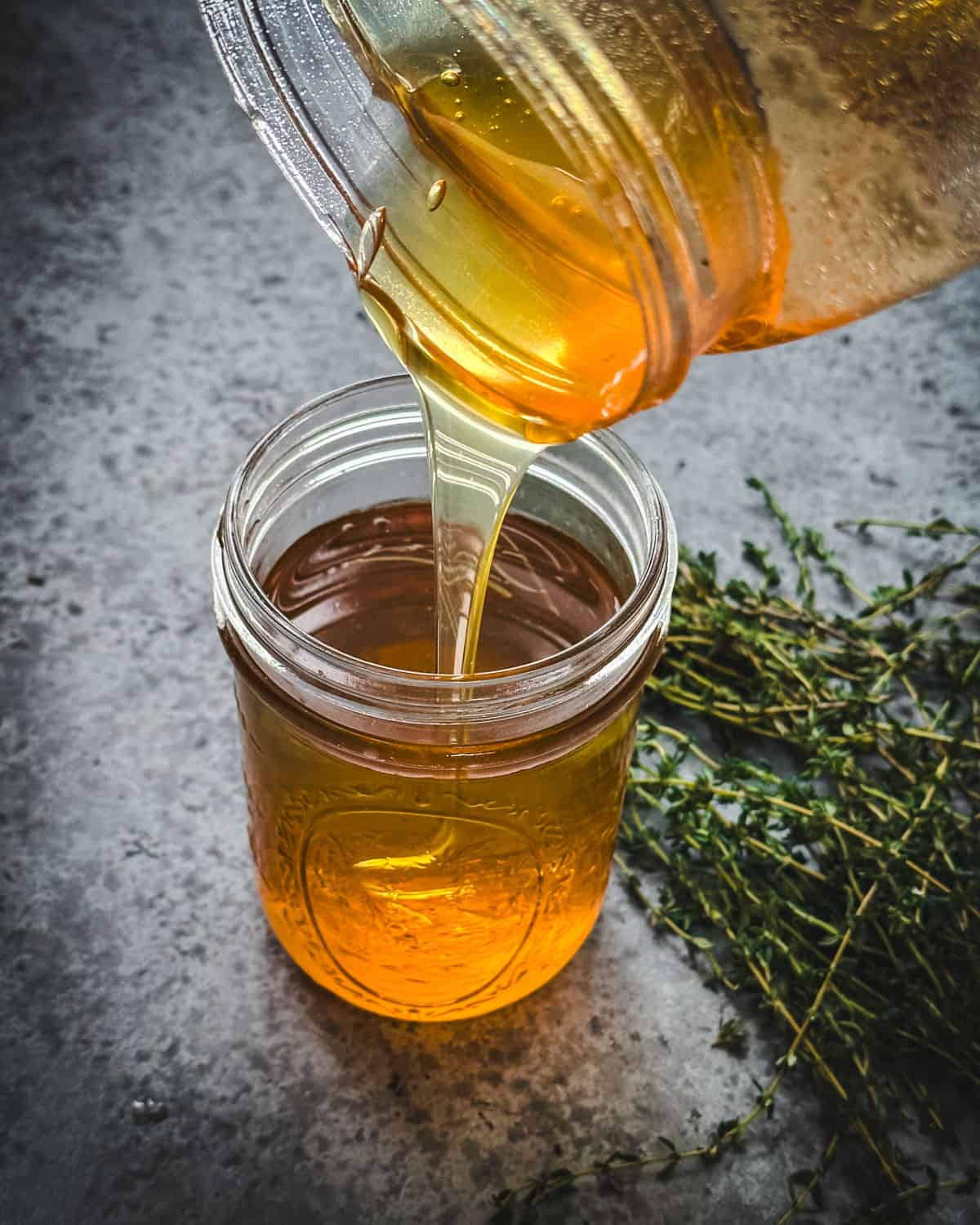 Honey pouring into the jar of thyme infusion, surrounded by fresh thyme sprigs. 