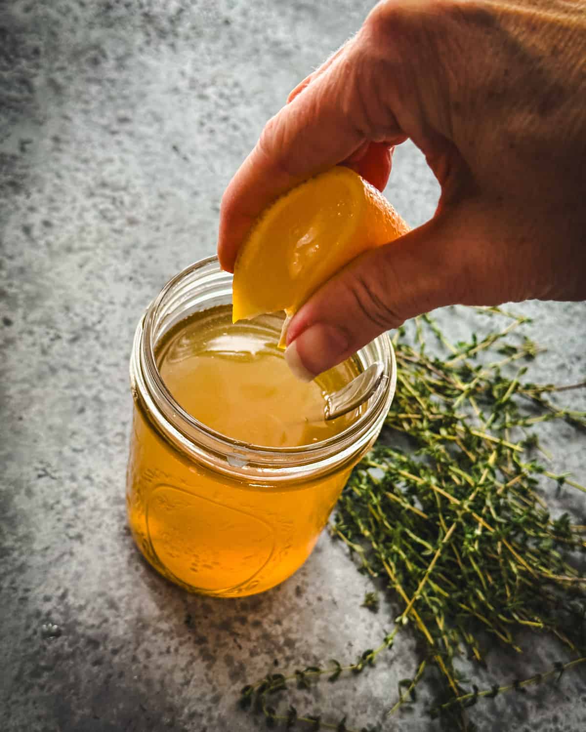 A hand squeezing a lemon wedge into a jar of thyme cough syrup, top view, with fresh thyme sprigs surrounding. 