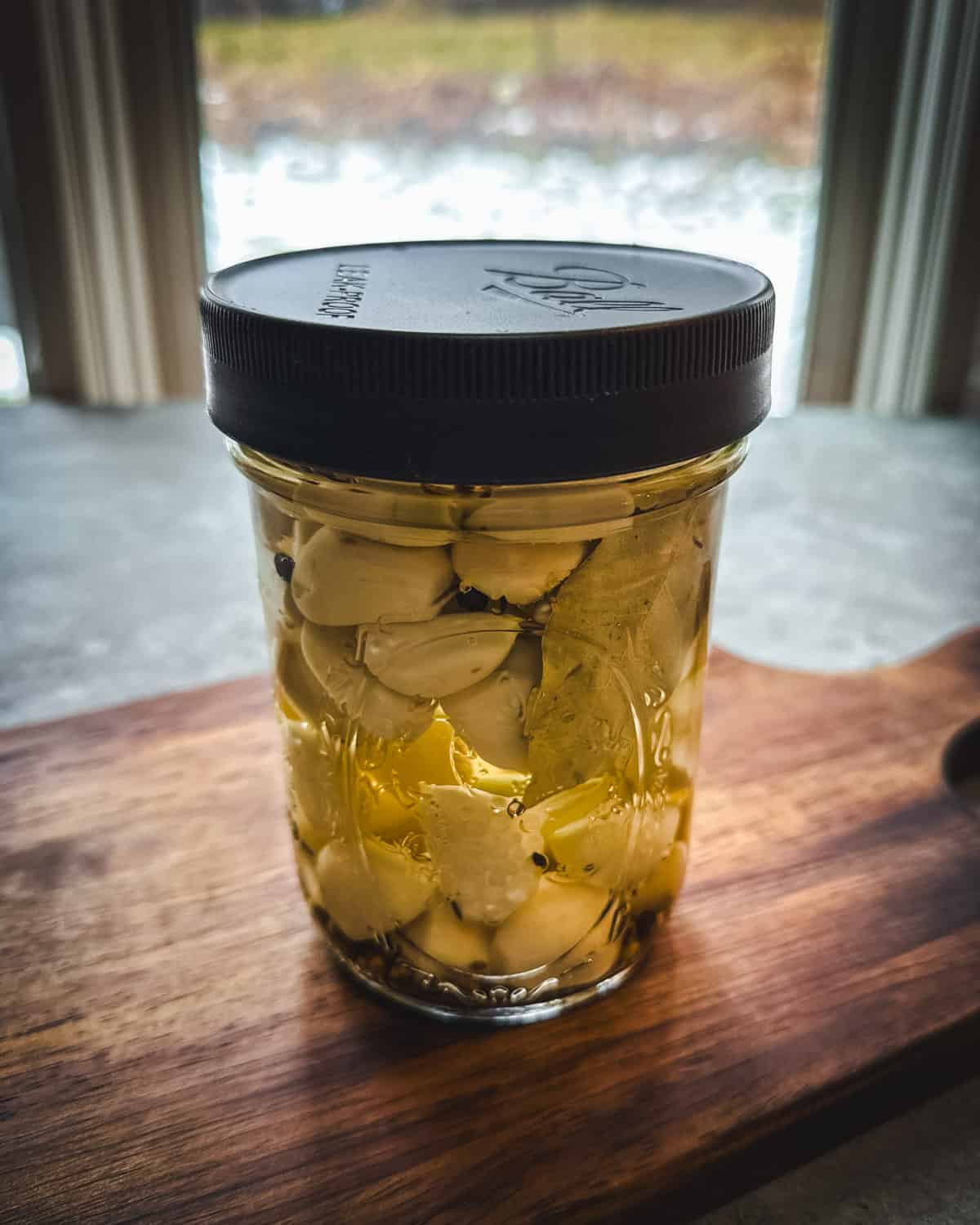 A jar of garlic in pickling brine, with a black cap on it, on a wood cutting board, with a window in the background. 